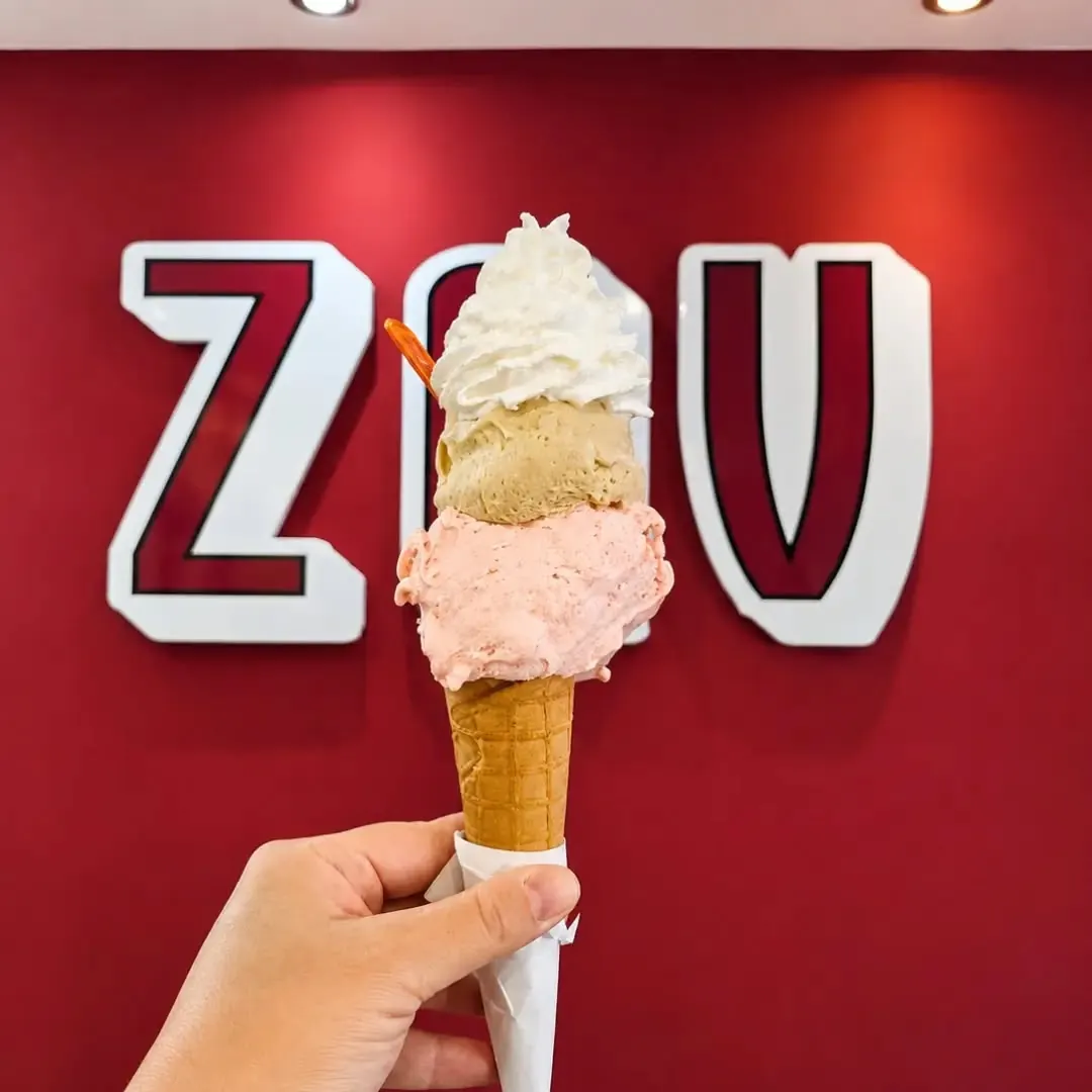 Un cono a tre gusti tenuto in mano davanti al muro bordeaux con le lettere ZAV del locale
